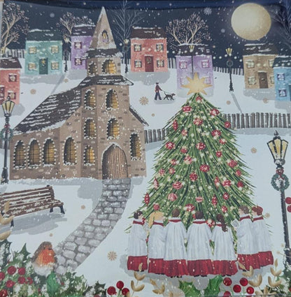 A square printed card or art print wrapped in clear plastic, placed on a light wood surface. The artwork depicts a snowy village scene with a Christmas tree, buildings, and a dark night sky.