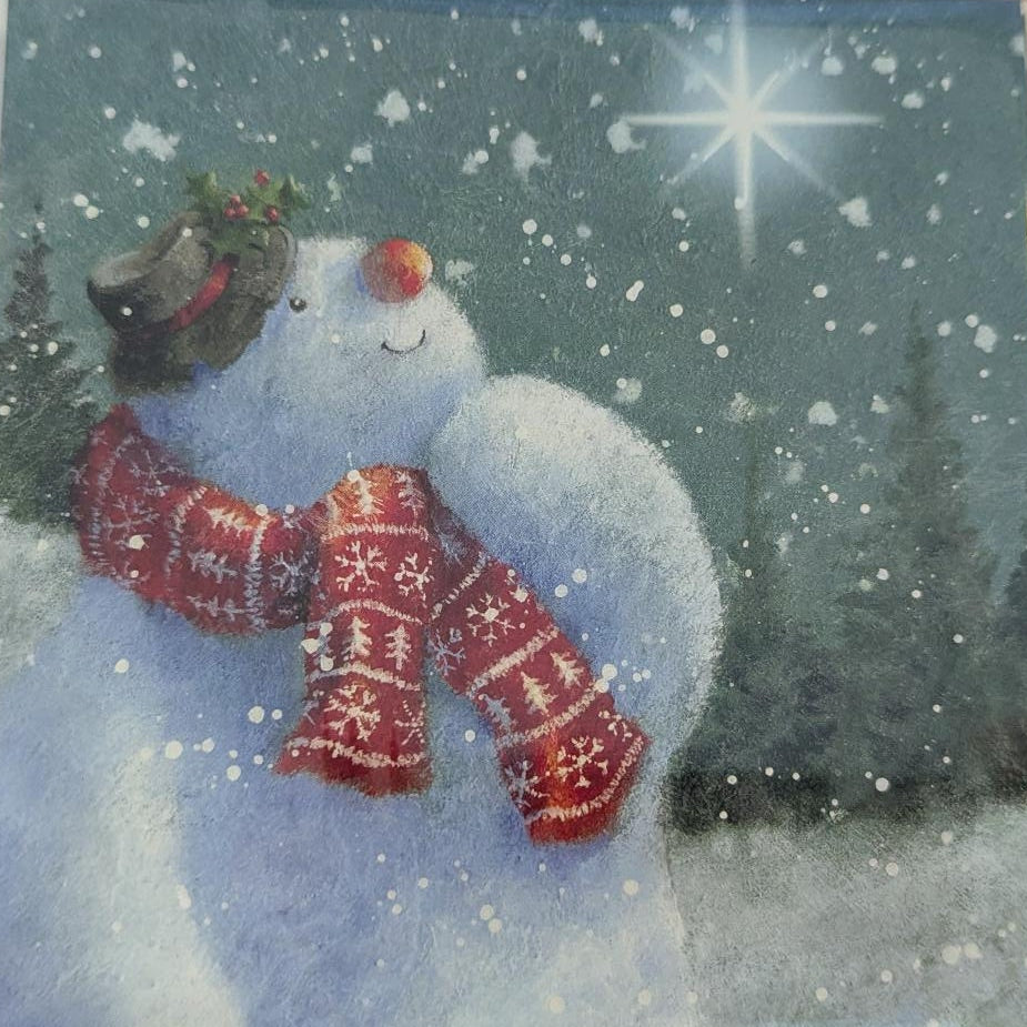 A square Christmas greeting card or art print in a clear plastic sleeve, featuring a snowman wearing a red patterned scarf and hat against a snowy blue-gray background, placed on a light wood surface.